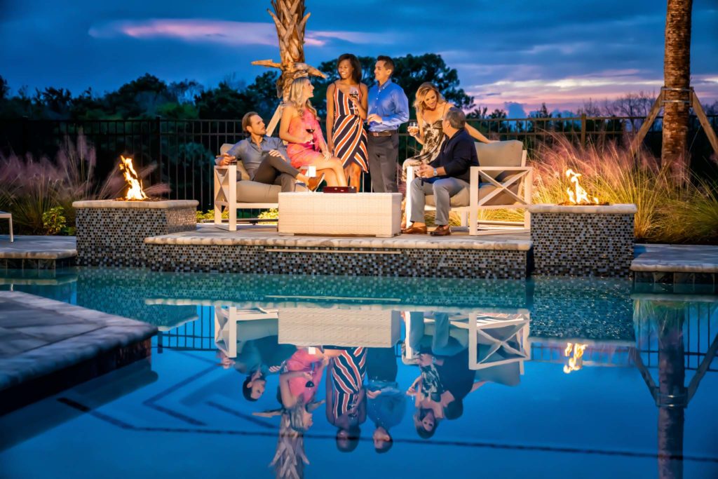 Group Of Three Couples Sitting At Night By The Spectrum Clubhouse Pool Next To A Pair Of Fire Pits.